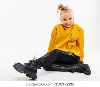 Pretty Blonde Little Girl Sitting In Studio Floor In A Leather Mustard Jacket, Black Snake Texture Leggings And Boots. High Fashion, Full Length Standing Pose, Isolated Against A Studio Background