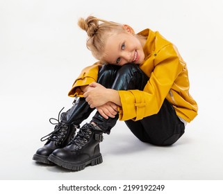 Pretty Blonde Little Girl Sitting In Studio Floor In A Leather Mustard Jacket, Black Snake Texture Leggings And Boots. High Fashion, Full Length Standing Pose, Isolated Against A Studio Background