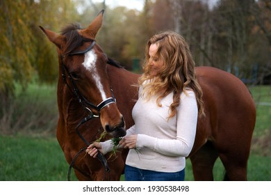 pretty, blonde, long-haired woman with many curls stands in the field and cuddles with her horse