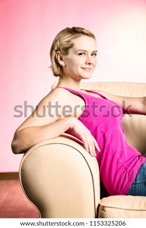 A pretty blonde teenage girl in a bright pink top sits on a sofa, feet up, leaning on the couch's arm.