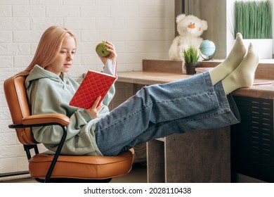 Pretty Blonde Teenage Girl In Home Clothes Reading Textbook,doing Homework In A Relaxed Pose At Home At Her Desk.Back To School Concept.