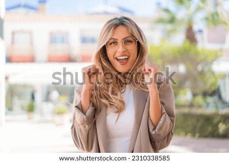 Pretty blonde Uruguayan woman with glasses at outdoors celebrating a victory in winner position