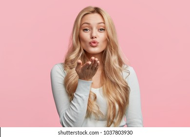 Pretty blonde woman blows passionate kiss into camera, expresses her love, isolated over pink background. Adorable young female model makes air kiss. People, body language and beauty concept