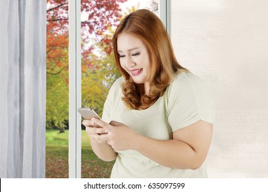 Pretty blonde woman with overweight body using cellphone at home and looks happy, shot with autumn background on the window