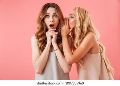 Pretty blonde woman in pajamas talking something to ear of shocked brunette woman over pink background