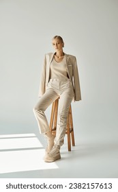 pretty blonde woman in pastel beige suit looking at camera on tall stool in sunshine on grey