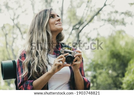 Pretty blonde woman standing in forest with camera and looking on scenery. Caucasian long-haired traveler walking or hiking in woods. Blurred background. Tourism, trip and summer vacation concept