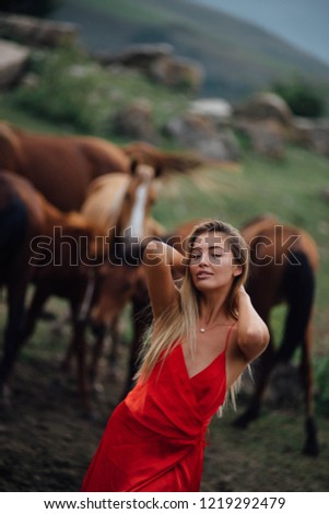 Pretty blonde woman walking near the horses in the red dress. Nature landscape around. Fresh air. Travel far away from cities.