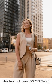 pretty blonde woman walking in sunny city street in beige suit with a purse in urban fashion style, smiling, summer trend season, wearing vest and trousers, glam hair hairstyle