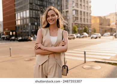 pretty blonde woman walking in sunny city street in beige suit with a purse in urban fashion style, smiling, summer trend season, wearing vest and trousers, glam hair hairstyle