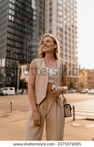 pretty blonde woman walking in sunny city street in beige suit with a purse in urban fashion style, smiling, summer trend season, wearing vest and trousers, glam hair hairstyle