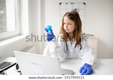 pretty blonde and young schoolgirl girl plays doctor and has white coat on and blue protective gloves and watches a blue ball, a model of a virus