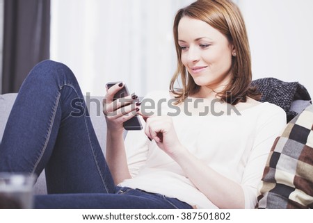 Pretty brown haired young woman dressed casual sitting on a sofa while playing with her smartphone