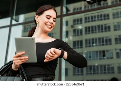Pretty brunette caucasian woman dressed leather suit with shoulder bag stand outside hold tablet pc, looking at swart wrist wireless watch fitness bracelet, checking time, waiting in a hurry, punctual