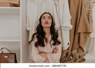 Pretty Brunette Curly Asian Woman With Red Lips In Pink Pink Pajamas Looks Up In Dressing Room. Sad Girl Poses In Wardrobe.