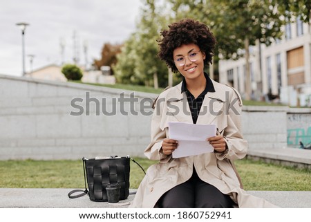 Pretty brunette curly woman in eyeglasses and beige trench coat holds paper sheet, smiles and sits outdoors.