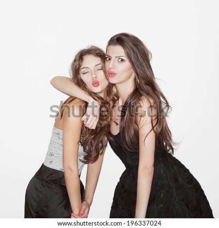 Pretty brunette girl friends having fun. Looking at camera and and showing kisses. One hugs another. Festive mood. Inside