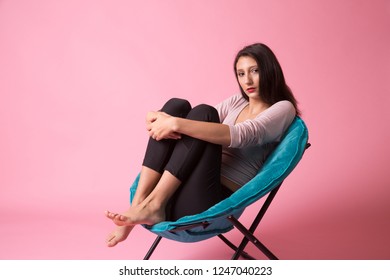 Pretty brunette teen girl sitting on a blue saucer chair on a pink background in teen lifestyle poses