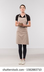 Pretty brunette waitress in workwear making notes in small notepad while standing in front of camera in isolation