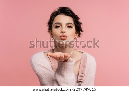 pretty brunette woman in cozy cardigan sending air kiss and looking at camera isolated on pink