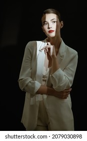 Pretty Brunette Woman With Evening Makeup Dressed In Elegant White Suit Poses In A Beam Of Light On A Black Background. Makeup And Cosmetics.