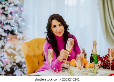 Pretty Brunette Woman With Long Hair In Bright Winter Sweater Posing With Gold Glass Of Champagne Sitting Near Christmas Table. Holiday Concept