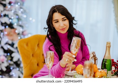 Pretty Brunette Woman With Long Hair In Bright Winter Sweater Posing With Gold Glass Of Champagne Sitting Near Christmas Table. Holiday Concept