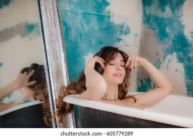 Pretty brunette woman taking a bath at art bathroom with old mirror and listen music in headphones