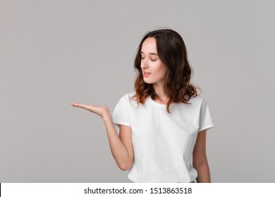 Pretty Brunette Woman In A White Tee Holding Palm Of A Hand With Empty Space For Product Placement Standing Isolated Over Grey Background. Copy Space Of Advertising.