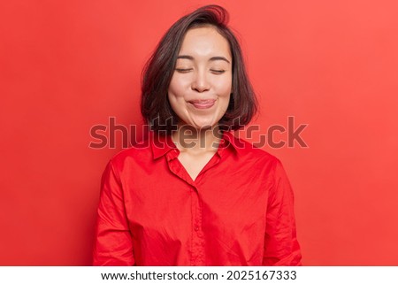 Pretty brunette young Asian woman closes eyes licks lipsfrom temptation to taste something delicious shows tongue imagines eating delicious food wears shirt isolated over vivid red background
