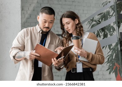 pretty businesswoman pointing at folder and discussing startup project with focused asian colleague - Powered by Shutterstock