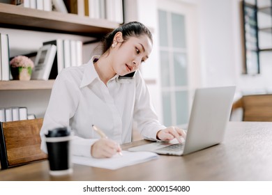 Pretty businesswoman sitting on the phone at her desk with laptop computer and notepad in hustle and bustle, rush hour work concept, working from the smart office.
