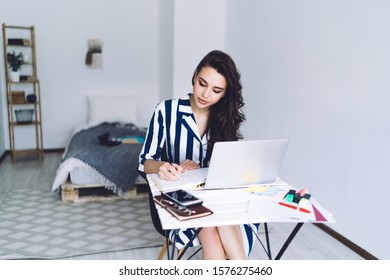 Pretty Calm Pensive Woman Taking Notes In Notepad While Sitting At Desk With Laptop Phone And Stationery In Light Bedroom With White Walls On Blurred Background