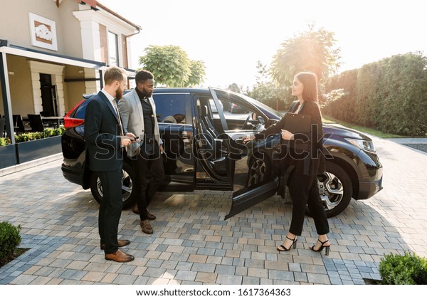 Pretty car saleswoman in business wear helping\
to young two multiethnical businessmen clients to make decision\
showing a new car, opening car doors, while standing on the yard of\
car salon outdoors