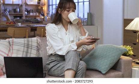 Pretty Career Woman Relaxing On Couch At Home With Hot Tea While Reading Online Article On Mobile Phone. Technology And Genuine Lifestyle