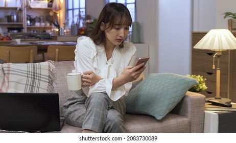 Pretty Career Woman Relaxing On Couch At Home With Hot Tea While Reading Online Article On Mobile Phone. Technology And Genuine Lifestyle