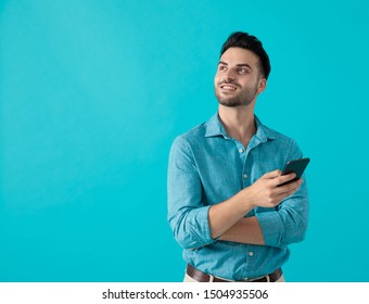 Pretty Casual Man In Blue Shirt Is Standing With Crossed Arms While Looking Up And Holding His Phone Curious On Blue Studio Background