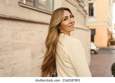 Pretty Caucasian Adult Lady With Blond Hair Looks Over Her Shoulder While Walking Down Street. Beauty Model Wear Casual Outerwear In Spring. Good Mood, Fashion Trends