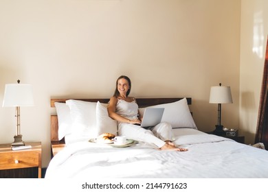Pretty Caucasian blogger with laptop technology smiling during leisure dayspa in hotel bedroom, cheerful female freelancer with digital netbook technology enjoying room service - yourself time