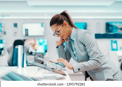 Pretty Caucasian Brunette With Ponytail Dressed In Formal Wear And With Eyeglasses Leaning On Counter And Trying Out Tablet. Tech Store Interior.