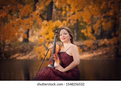 pretty Caucasian female violinist in long burgundy elegant dress sitting near water with reflection of autumnal yellow leaves in park in sunny fall day 