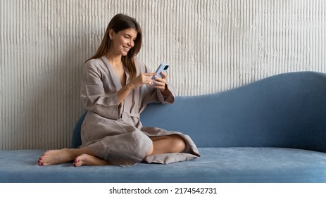 Pretty Caucasian Woman Using Smartphone Sitting On Blue Sofa , Having Rest In Home Dressing Gown, Watching Funny Videos, Stories, Browsing Social Media