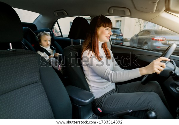 Pretty Caucasian woman, young mother driving a\
car, having her little baby girl in a child seat behind. Safety\
driving, children car seat\
concept