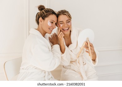 Pretty caucasian young women wear bathrobes admire themselves looking in mirror on white background. Blondes use eye patches for dark circles. Beauty and personal care, skin and face care concept - Powered by Shutterstock