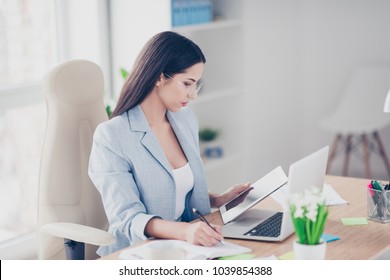 Pretty, charming, clever, smart, brunette boss wearing jacket and glasses making notes in notebook, looking at gadget, sitting on chair at desk in work station, organize her working day