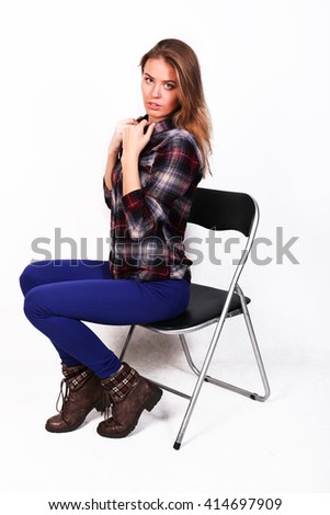 pretty charming girl Caucasian blonde in a plaid shirt and blue pants sitting on a chair, closing collar and chest by hands, modest, isolated on white