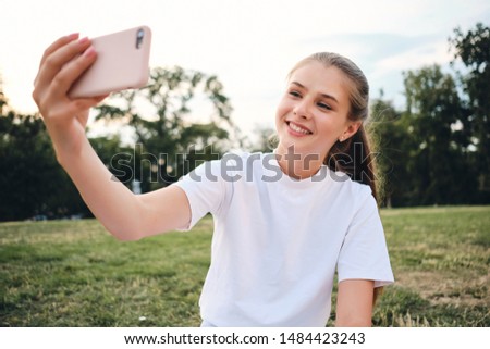 Pretty cheerful girl in white T-shirt happily taking photo on cellphone sitting on lawn in city park