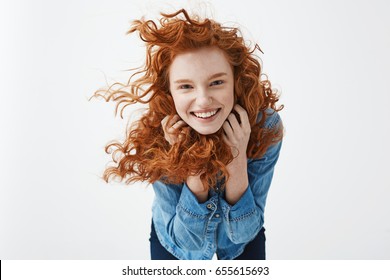 Pretty cheerful redhead girl with flying curly hair smiling laughing looking at camera over white background.