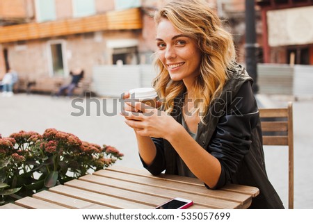 Pretty cheerful young blonde girl in smart casual eclectic outfit sits on cafe terrace while drinking coffee from paper cup. Coffee to go