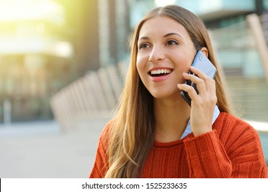 Pretty cheerful young woman talking on phone and looks straight ahead. Girl holding smartphone close to ear standing alone outside. Daylight with modern cityscape on the background.
